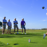 A view of six people at a golf course and one hitting the shot