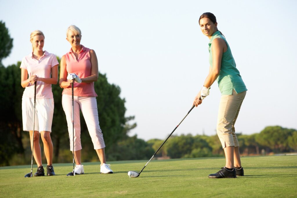 Three women standing at a golf course and one hitting the golf ball