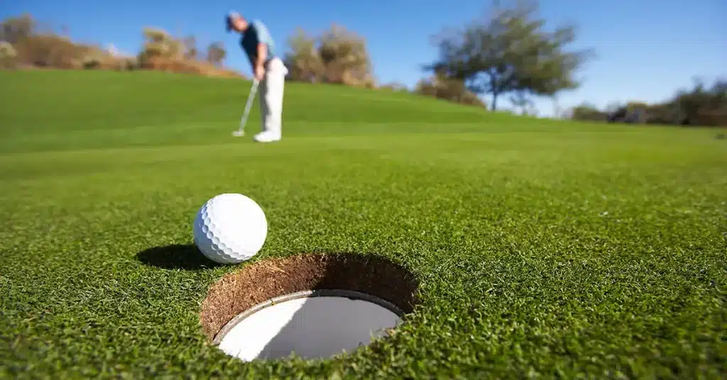 zoomed in view of a golf hole a ball placed near it blurred view of a golfer holding club